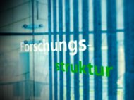 Grüne und weiße Schrift "Forschungsstruktur" vor Glaswand mit Streifenmuster.