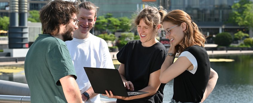 Vier Personen stehen im Freien und diskutieren, während sie auf einen Laptop schauen, umgeben von modernem Stadtambiente.