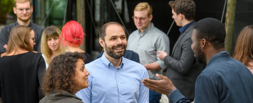 Mehrere Personen stehen draußen zusammen und unterhalten sich. Im Vordergrund sticht eine Person mit roten Haaren hervor, während die anderen leger gekleidet sind, was auf eine entspannte oder gesellige Atmosphäre hindeutet.