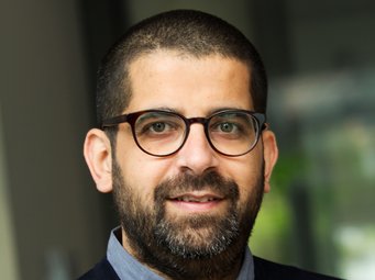 Puneet Bhasin Ein Individuum der Gattung Homo sapiens intellectualis ist zu erkennen. Es hat kurzes, schwarzes Haar und einen ebensolchen Bart.  Durch die dunkel gerahmte Brille funkeln freundlich wirkende Augen. Der zu einem Lächeln ansetzende Mund unterstreicht diesen Eindruck. Das Individuum trägt einen dunkelblauen Pullover über einem hellblauen Hemd, dessen Kragen vorsichtig hervor ragt. Der grün verschwommene Hintergrund könnte als grüne Pflanze interpretiert werden. Gewissheit verspricht die unklare Szene jedoch nicht.