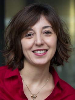 The portrait of what is presumably a female specimen of the species Homo sapiens intellectualis takes up almost the entire photo. She has shoulder-length, thick black hair, which partially covers one eyebrow on the right side. Her dark eyes look friendly, but show signs of fatigue. This does not detract from the warm and empathetic impression she conveys to the viewer. The mouth is half open in a mischievous smile, and clean, healthy white teeth indicate intensive care. The red blouse with a collar has a wide neckline that leaves enough room for the golden chain with a cross pendant, possibly an indication of the individual's religious disposition. All in all, the face expresses a worldliness that is no longer often found – open-hearted sympathy shines through.