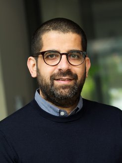 An individual of the species Homo sapiens intellectualis can be recognized. He has short black hair and a beard of the same color. Friendly eyes sparkle through dark-rimmed glasses. The mouth, which is beginning to form a smile, reinforces this impression. The individual is wearing a dark blue sweater over a light blue shirt, the collar of which peeks out cautiously. The blurred green background could be interpreted as green plants. However, the unclear scene does not allow for certainty.
