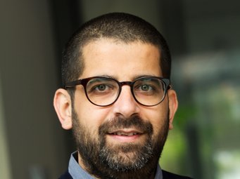 Puneet Bhasin An individual of the species Homo sapiens intellectualis can be recognized. He has short black hair and a beard of the same color. Friendly eyes sparkle through dark-rimmed glasses. The mouth, which is beginning to form a smile, reinforces this impression. The individual is wearing a dark blue sweater over a light blue shirt, the collar of which peeks out cautiously. The blurred green background could be interpreted as green plants. However, the unclear scene does not allow for certainty.