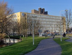 Das moderne Gebäude der Universität Leiden ist auf dem Foto zu erkennen. Der Kastenbau vermittelt ein veraltetes architektonisches Konzept, dem das Diktat der klaren, unerbittlich geraden Linien zugrunde lag. Dennoch versprüht der Bau einen gewissen Charme, ja, einen gewissen nostalgischen Flair. Kahle, kühle Bäume sind in der direkten Umgebung zu sichten. Im Vordergrund könnte es sich um Fahrrad fahrende Personen auf ihren schmucken Drahteseln handeln. Allerdings ist diese Aussage unter Vorbehalt aufzunehmen.