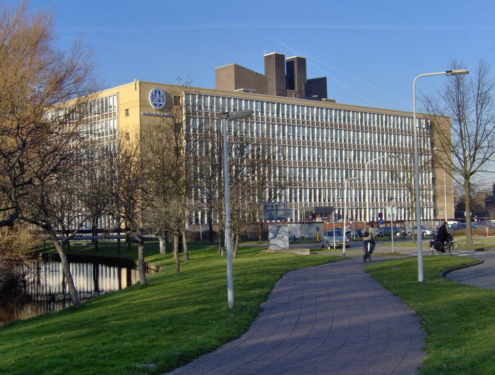 Das moderne Gebäude der Universität Leiden ist auf dem Foto zu erkennen. Der Kastenbau vermittelt ein veraltetes architektonisches Konzept, dem das Diktat der klaren, unerbittlich geraden Linien zugrunde lag. Dennoch versprüht der Bau einen gewissen Charme, ja, einen gewissen nostalgischen Flair. Kahle, kühle Bäume sind in der direkten Umgebung zu sichten. Im Vordergrund könnte es sich um Fahrrad fahrende Personen auf ihren schmucken Drahteseln handeln. Allerdings ist diese Aussage unter Vorbehalt aufzunehmen.