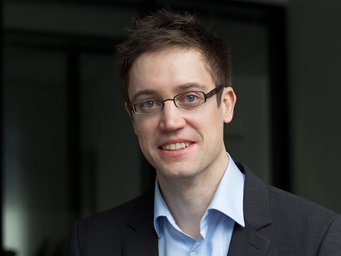 Man with short, dark hair wearing a dark suit and light blue shirt, standing in front of a blurry, dark background.