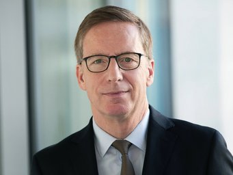 Das Porträtfoto zeigt Michael Hüther, Vorsitzender des Kuratoriums des MPIfG. Der mittelalterliche Mann trägt Anzug und Krawatte sowie ein hellblaues Hemd. Das kurze volle Haar ist nach rechts gekämmt, durch eine Brille schaut er freundlich lächelnd dem Betrachter entgegen.