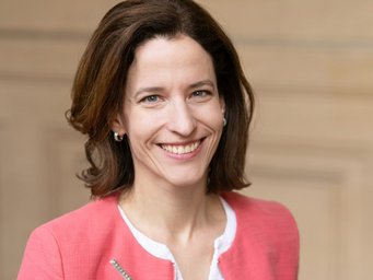 Woman with brown shoulder-length hair, wearing a coral pink blazer over a white top, accessorized with small hoop earrings.