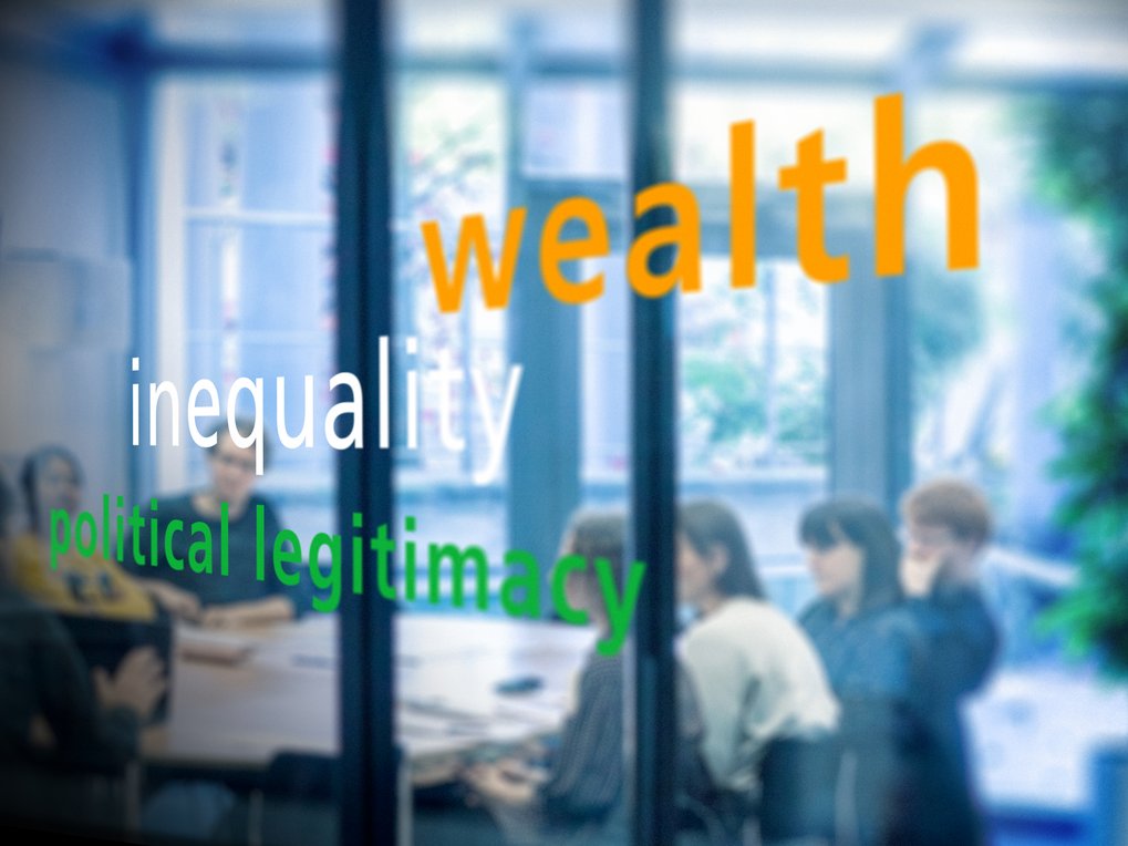A blurred image showing the words "wealth," "inequality," and "political legitimacy" on a glass surface, with people seated around a table in a meeting setting.