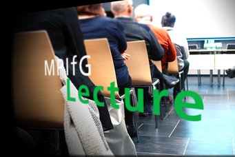 Attendees sitting in rows of wooden chairs in a lecture setting, with a glass wall reflecting the text "MPIfG Lecture" prominently in green.