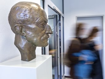 A bronze bust displayed on a white pedestal in a hallway, with several individuals walking past in the background, creating a sense of motion.