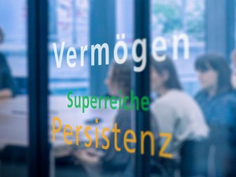 Office glass wall featuring words "Vermögen" in white, "Superreiche" in green, and "Persistenz" in orange, with seated individuals in the background.