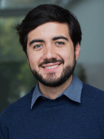 An individual with dark hair is dressed in a blue sweater layered over a striped collared shirt, standing in front of a blurred background.