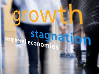 Text displayed on a glass surface: "growth stagnation peripheral economies" with blurred figures walking in the background.