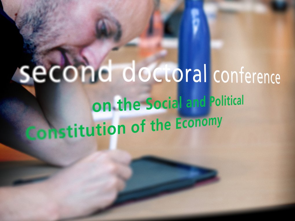 A person is writing on a tablet during the "second doctoral conference on the Social and Political Constitution of the Economy." A blue water bottle is also visible on the table.