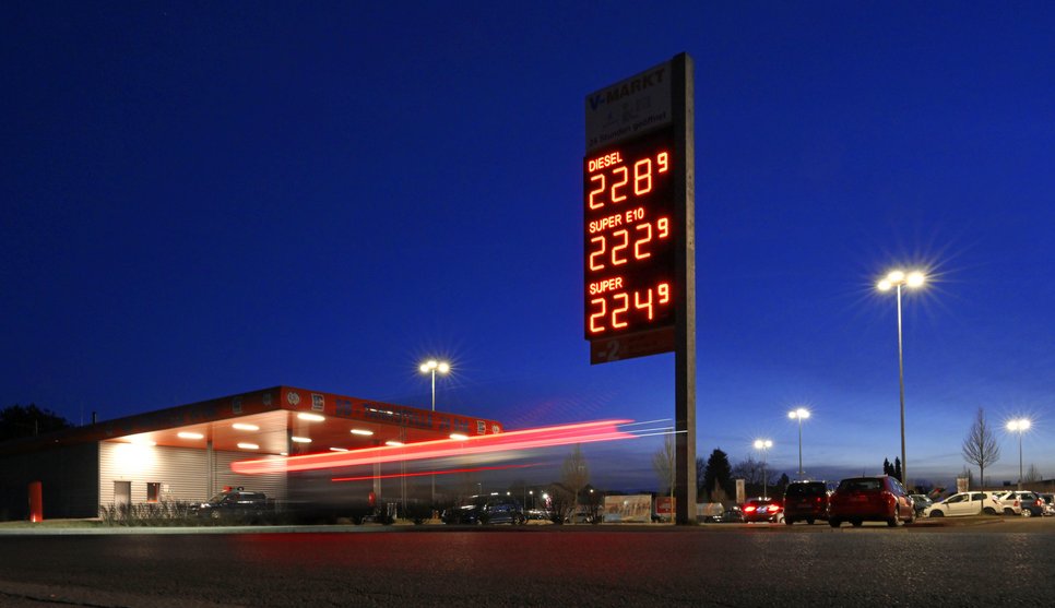 Tankstelle bei Nacht mit beleuchteter Preisanzeige für Kraftstoffe, darunter Diesel, Super E10 und Super, umgeben von parkenden Autos.