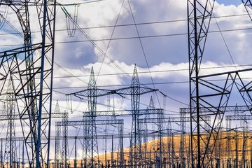 Ein Netz aus Hochspannungsmasten und Drähten erstreckt sich vor einem bewölkten Himmel, während im Hintergrund ein sanfter Hügel sichtbar ist.