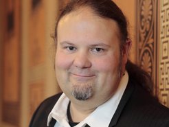 Mann mit schwarzem Anzug und weißem Hemd steht vor einer Wand mit komplexen, braunen Mustern und Verzierungen.