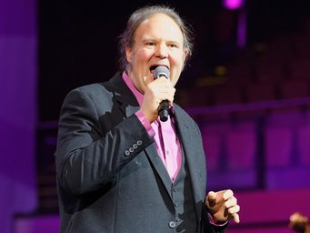 Wissenschaftler oder Musiker? Lange blieb für den Juniorprofessor auf befristeter Stelle offen, welches seiner beiden herausragenden Talente den Beruf prägen würde. Person in einem eleganten grauen Anzug mit rosa Hemd hält ein Mikrofon, steht im Fokus von violettem Licht auf einer Bühne.