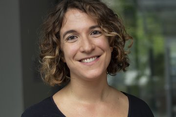 Person mit schulterlangem, lockigem Haar trägt ein schlichtes schwarzes Oberteil. Der Hintergrund ist leicht verschwommen.