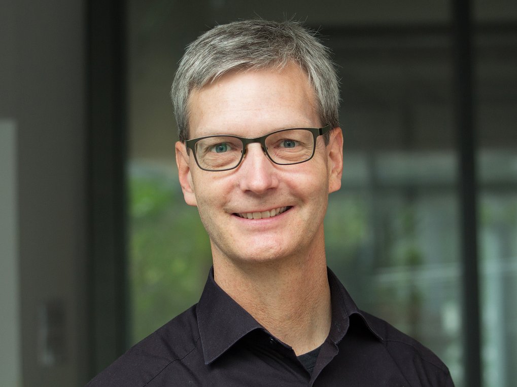 Ein Mann mit grauem Haar trägt ein schwarzes Hemd. Der Hintergrund ist unscharf, mit einem Hauch von Grün und strukturellen Details.