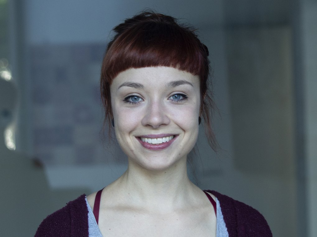 Eine Person steht in einem Innenraum, trägt ein graues Top mit einem bordeauxroten Pullover, Haare zu einem lockeren Dutt gebunden.