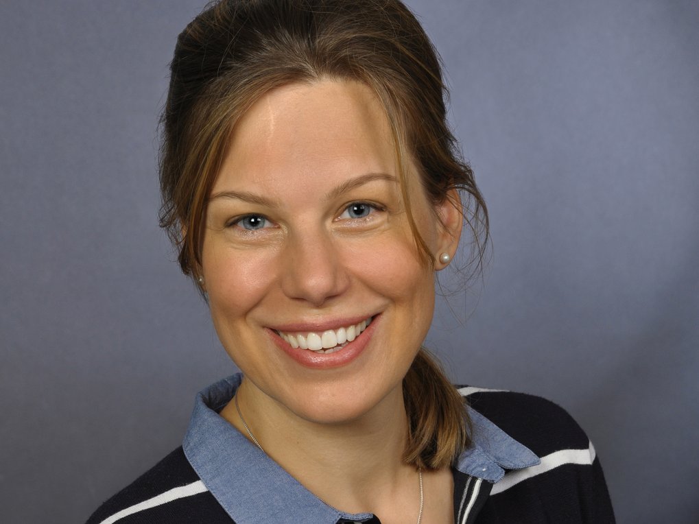 Frau mit braunen Haaren, trägt Perlenohrring und gestreiftes Oberteil. Der Hintergrund ist einfarbig grau.