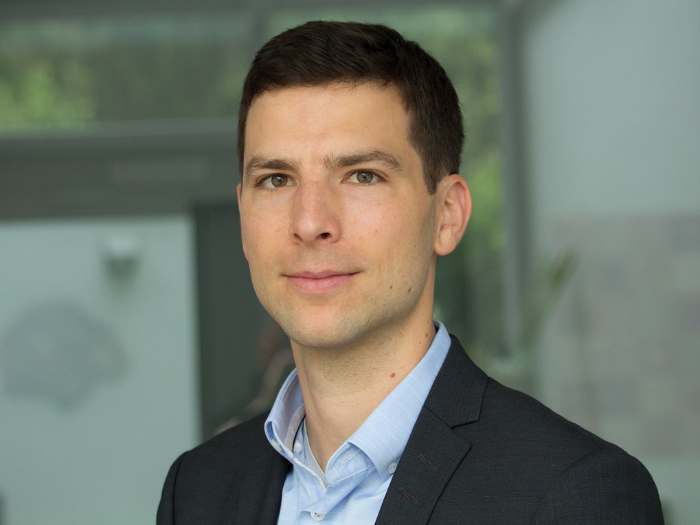 Ein Mann in einem dunklen Anzug und einem hellblauen Hemd steht vor einem Hintergrund, der durch Fenster und Pflanzen geprägt ist.