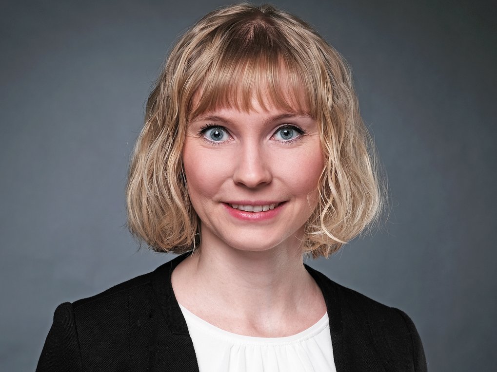 Blondhaarige Person trägt einen schwarzen Blazer und eine weiße Bluse vor einem grauen Hintergrund.