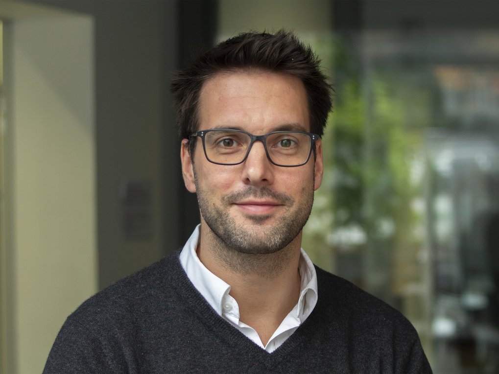 Individuum trägt einen dunkelgrauen Pullover über einem weißen Hemd, im Hintergrund sind unscharfe Büroumgebungen erkennbar.