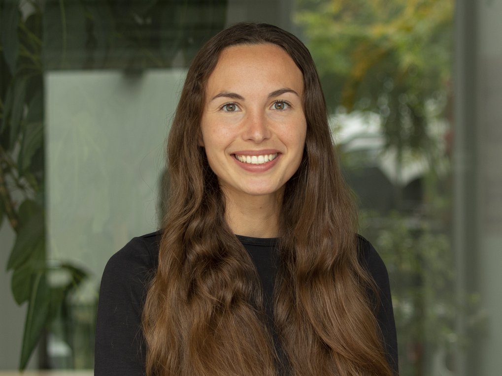 Eine Person mit langen, braunen, gewellten Haaren steht vor einem Fenster. Im Hintergrund sind unscharf Pflanzen und Bäume zu erkennen.