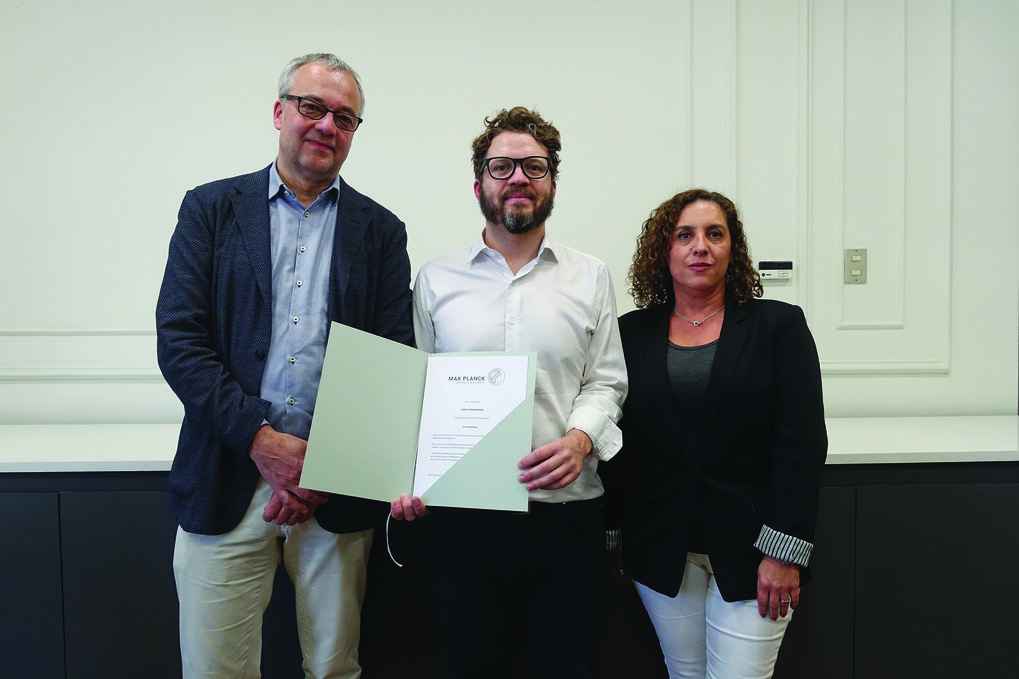 Jens Beckert, Aldo Madariaga, and Rossana Castiglioni (Dean of the Faculty of Social Sciences and History of the University Diego Portales) at the presentation of the letter of appointment Three people pose indoors, the person in the middle displays a certificate from the Max Planck Institute, suggesting an award or achievement.