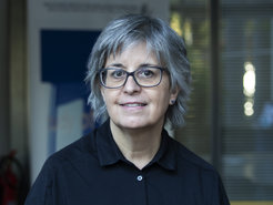 Freundlich dreinblickende Person, halblanges Haar, Brille, schwarzes Hemd vor verschwommenem Hintergrund aufgenommen.