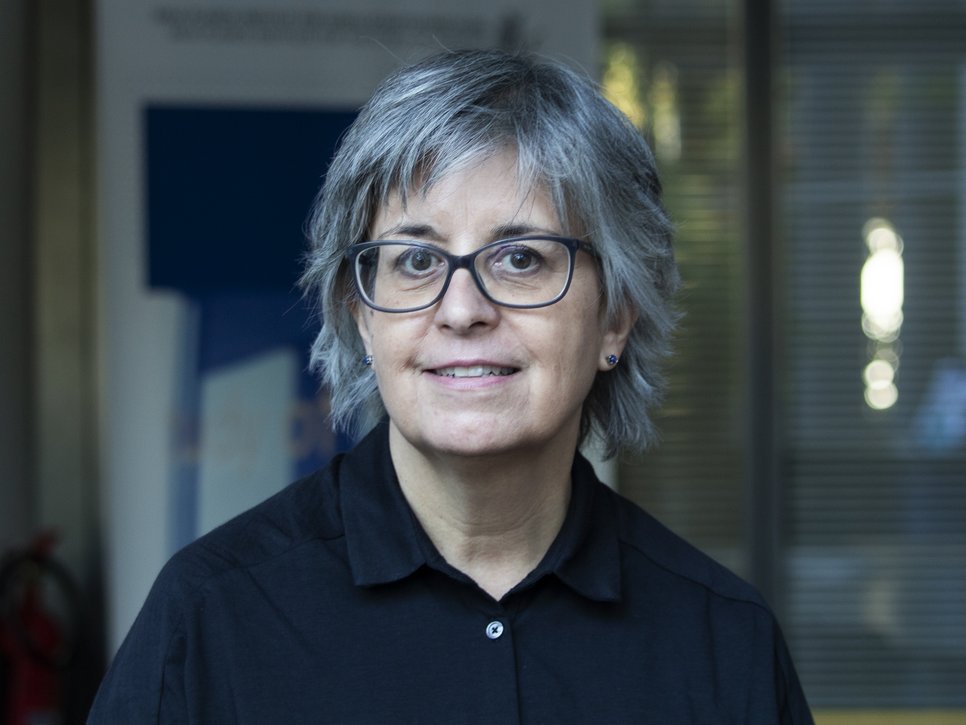 An individual with shoulder-length gray hair and a black button-up shirt is indoors, with a background that includes blurred architectural and interior elements.