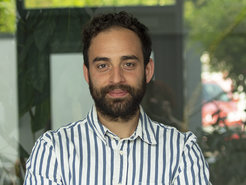 A person in a blue and white striped shirt is positioned indoors, with large leafy plants and a glass wall creating a modern backdrop.