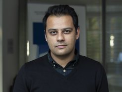 Person trägt dunkelblauen Pullover über einem karierten Hemd, steht in einem Büro mit Glasfenstern und unscharfem Hintergrund.