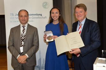 Das Foto zeigt die Szene anlässlich der Vergabe der Otto-Hahn-Medaille an Hannah Pool. Pool in der Mitte die Medaille zeigend, rechts und links zu ihrer Seite zwei Individuen männlichen Geschlechts, das eine mit zum Bund hin verschränkten Händen, das andere, auf der linken Seite Pools, hält die Urkunde aufgeschlagen in die Szene.