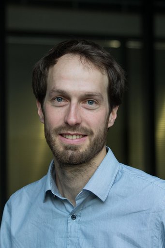 Das Foto zeigt ein junges menschliches Individuum männlichen Geschlechts. Das dunklelblonde Haar ist kurz geschnitten, ein ebensolcher Bart schmückt das freundlich lächelnde Antlitz. Es trägt ein hellbalues Hemd mit offenem Kragen. Der dunkle Hintergrund ist nicht definierbar.