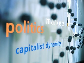 A reflective surface showcases the words "politics" in orange, "markets" in white, and "capitalist dynamics" in blue, with a blurred background.