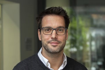 Das Foto zeigt ein junges menschliches Individuum männlichen Geschlechts. Das dunkle volle Haar ist kurz geschnitten, das freundlich lächelnde Antlitz ist mit einem Sehwerkzeug bestückt. Aus dem zum Hals hin offenen dunklen Pullover ragt der offene Kragen eines weißen Hemdes. Der diffus-farbige Hintergrund ist arg verschwommen und lässt sich kaum beschreiben.