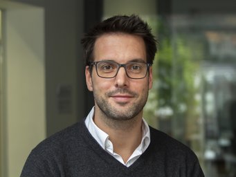 Georg Rilinger The photo shows a young male human individual. His dark, thick hair is cut short, and his friendly, smiling face is equipped with a visual aid. The open collar of a white shirt protrudes from the dark sweater, which is open at the neck. The diffuse-colored background is very blurred and difficult to describe.
