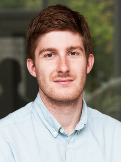 Auf dem Foto ist ein junges menschliches Individuum männlichen Geschlechts zu erkennen. Das dunkle volle Haar ist kurz geschnitten, das Gesicht lässt einen Bartanflug vermuten. Das zaghaft-freundlich lächelnde Antlitz deutet auf einen in sich ruhenden Charakter. Die helle Farbe des Hemdes samt Kragen unterstützt diesen Eindruck.