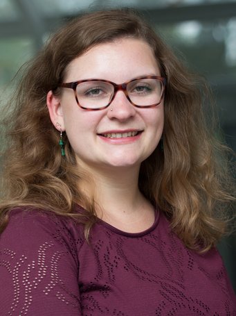 Das Foto zeigt ein junges, vermutlich weibliches menschliches Individuum, das freundlich lächelnd dem Betrachter entgegensieht. Das volle lange Haar ist zur rechten und zur linken Seite nach vorn gekämmt und verdeckt an dieser Stelle den weinroten Pullover, der trefflich zu den blauen Augen passt. Die Brille ist recht groß, sie dürfte ihren Zweck erfüllen.