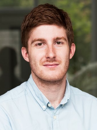 Eine Person trägt ein hellblaues Hemd. Im Hintergrund sind verschwommene Fenster und Pflanzen sichtbar, die einen dezenten, grünen Farbton hinzufügen.