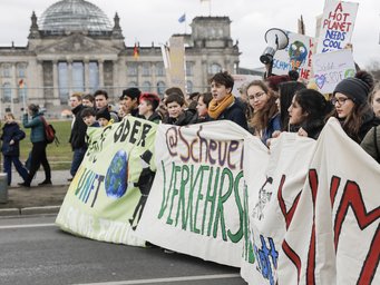 Das Foto zeigt Protestierende, die ein Banner mit Umweltbotschaften vor dem Reichstag vor sich her tragen. Schilder mit Aufschriften zu Klimaschutz und Verkehrswende sind sichtbar, im Hintergrund leicht verschwommen das Reichstagsgebäude in Berlin.