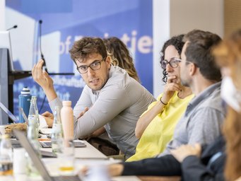 A group of conference attendees seated at a table with laptops, water bottles, and papers, actively participating in a discussion. The setting suggests a professional or academic environment.