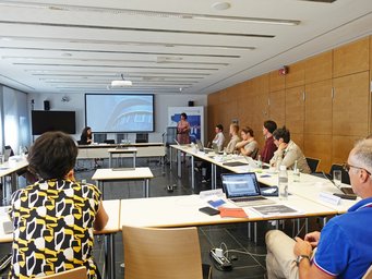 In a modern conference room, participants are seated around a U-shaped table, attentively listening to a speaker at a podium. A large screen displays a presentation, and various documents and laptops are visible on the tables.