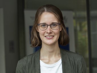 Auf dem Foto ist ein Mensch vermutlich weiblichen Geschlechts zu erkennen. Er hat schulterlanges braunes Haar und trägt einen grünen Blazer über einem weißen T-Shirt. Es steht vor einem unscharfen, nicht definierbarem Hintergrund, vermutlich einem Plakat vor einem Fenster.