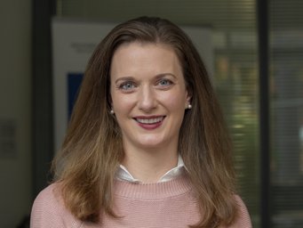 Person with shoulder-length brown hair, wearing a pink sweater and white shirt. Background with blurred office elements.