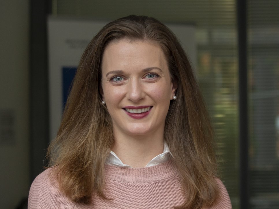 Person with shoulder-length brown hair, wearing a pink sweater and white shirt. Background with blurred office elements.